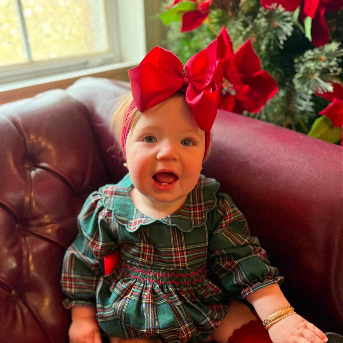 A baby girl wearing a festive holiday outfit, featuring a green and red plaid dress, red knee-high socks, and a large red bow headband, sitting on a dark red leather tufted couch.