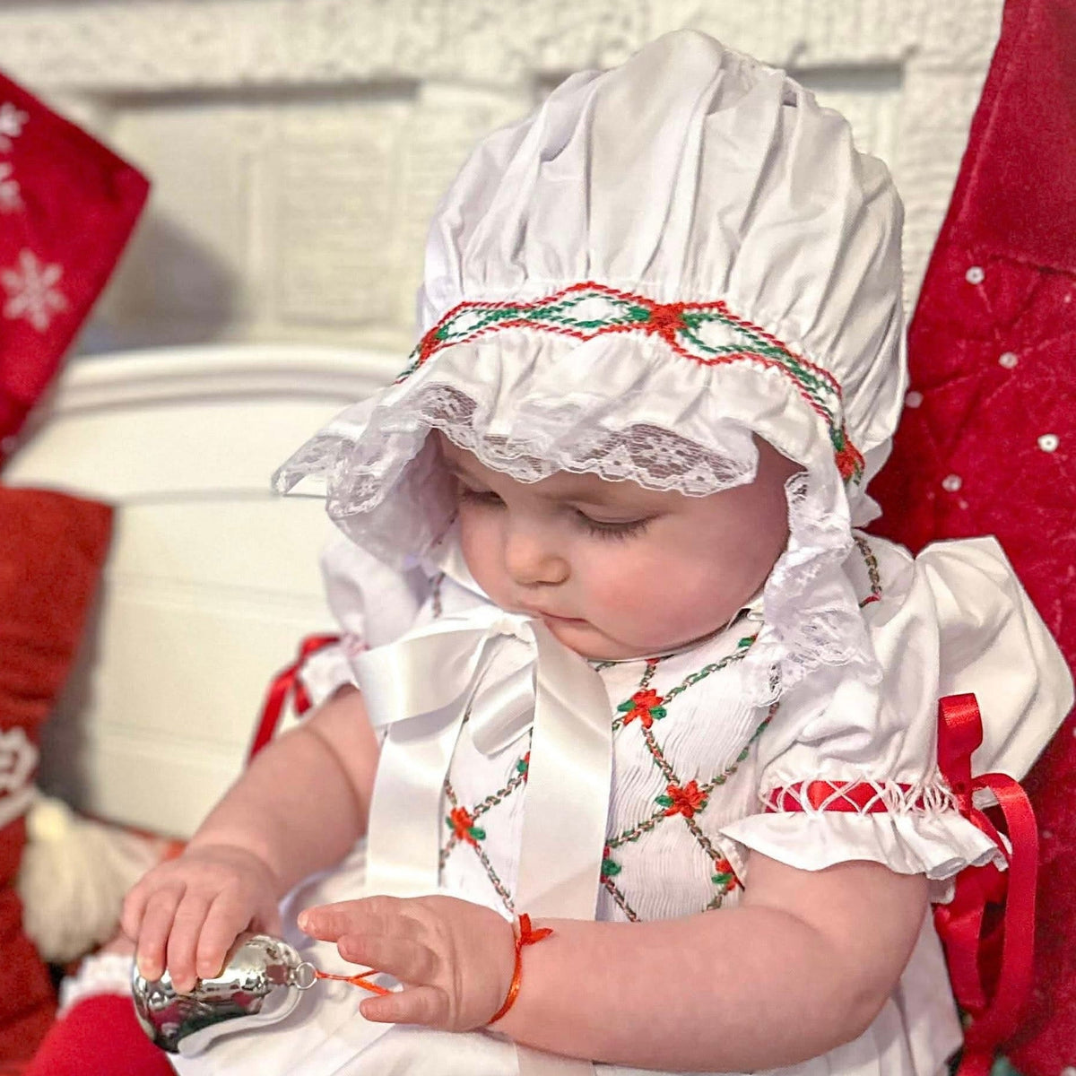 A baby wearing a white Christmas bonnet and dress with red accents, sitting on a plaid blanket and holding a small silver toy. Red holiday stockings and pillows are visible in the background.