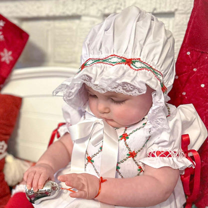 A baby wearing a white Christmas bonnet and dress with red accents, sitting on a plaid blanket and holding a small silver toy. Red holiday stockings and pillows are visible in the background.