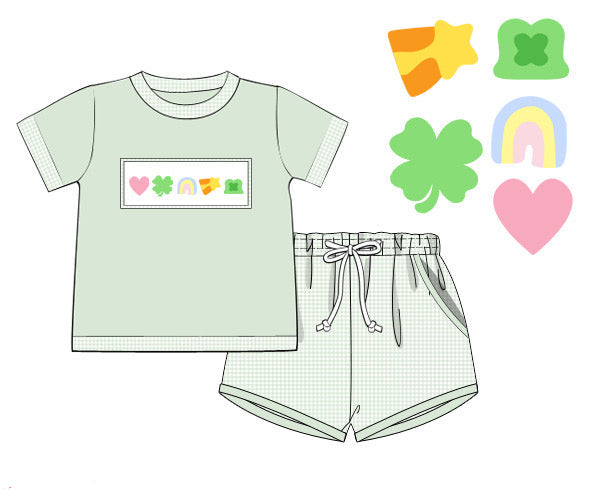 A flat product mockup showing a light sage green short-sleeve knit top and matching shorts set. The top features a rectangular chest graphic with pastel-colored shamrocks, a heart, a slice of cheese, and a small rainbow.