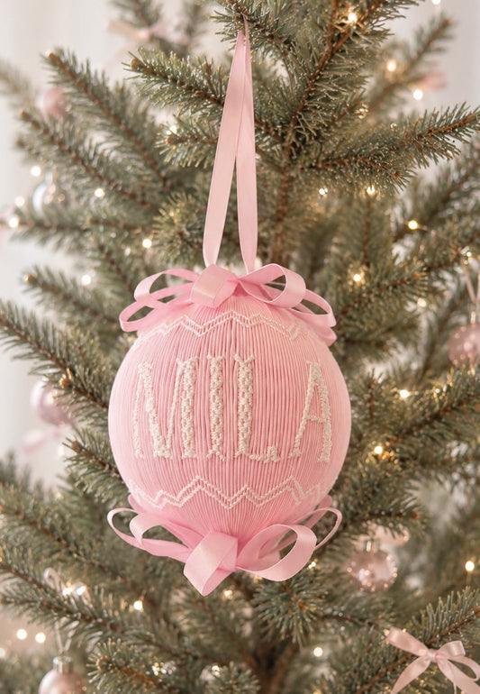 A pink personalized Christmas ornament hanging on a decorated Christmas tree. The ornament is a round pink fabric ball with white embroidered lettering that reads 'MILA' and pink ribbon bows at the top and bottom.