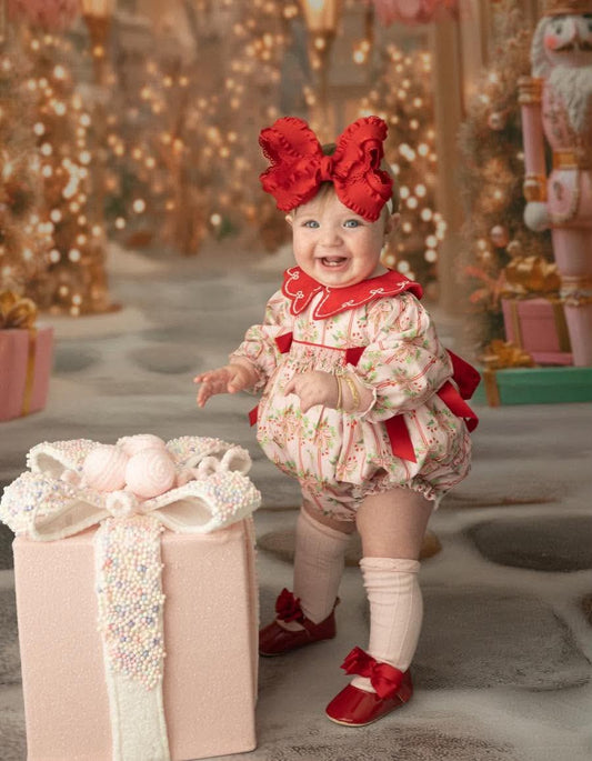 A baby wearing a festive holiday outfit with a red bow headband, a cream floral romper, red shoes, and white socks, standing next to a large pink gift box in a decorated Christmas-themed studio setting.